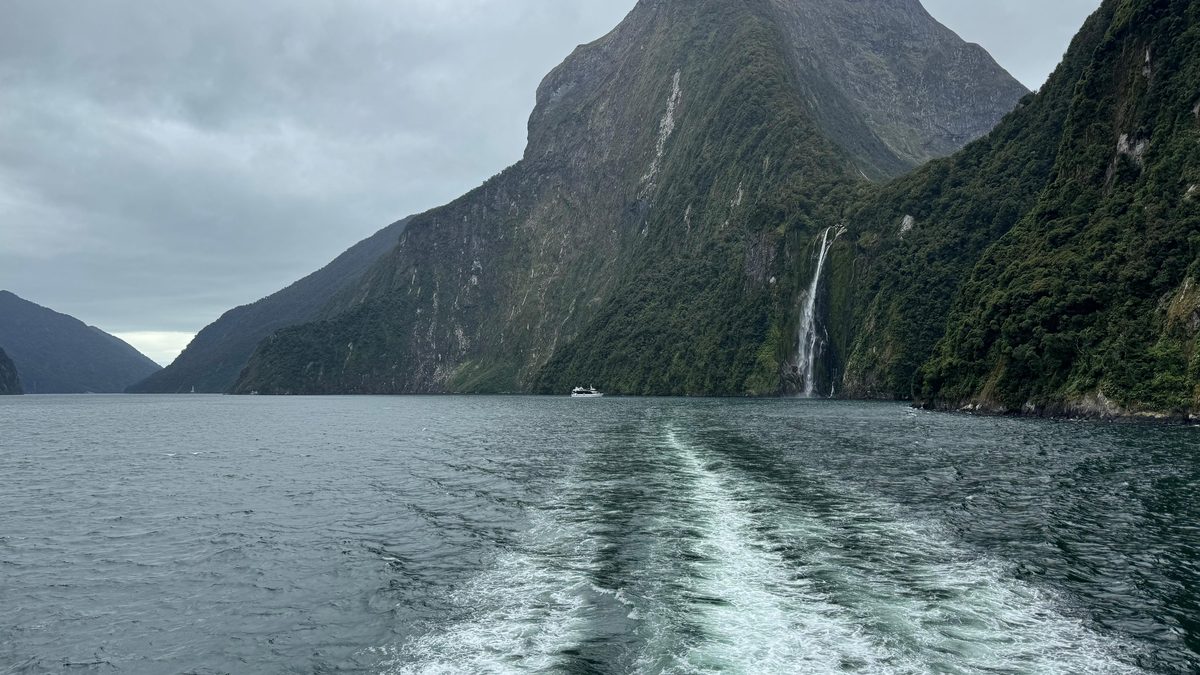 A steep fiord with waterfalls and a boat wake on dark green water.
