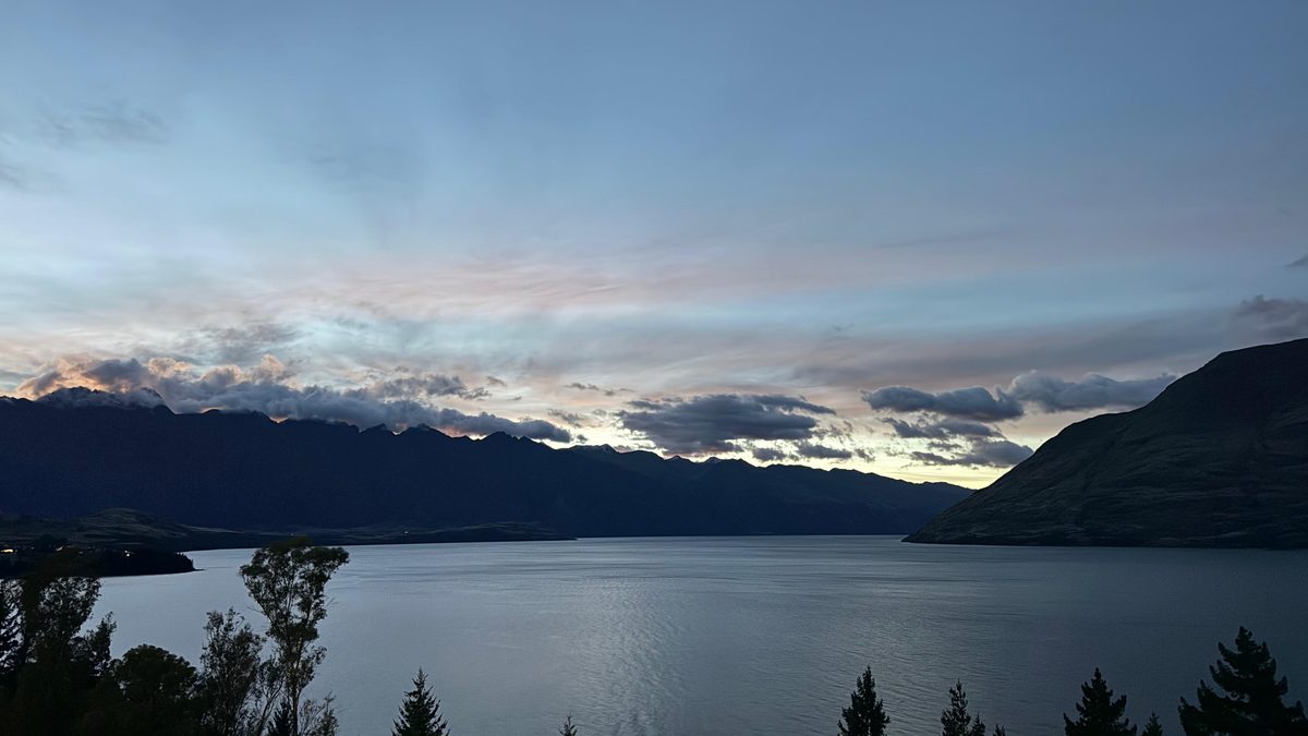 A lake at dusk with dark hills on both sides and pine trees in the foreground.