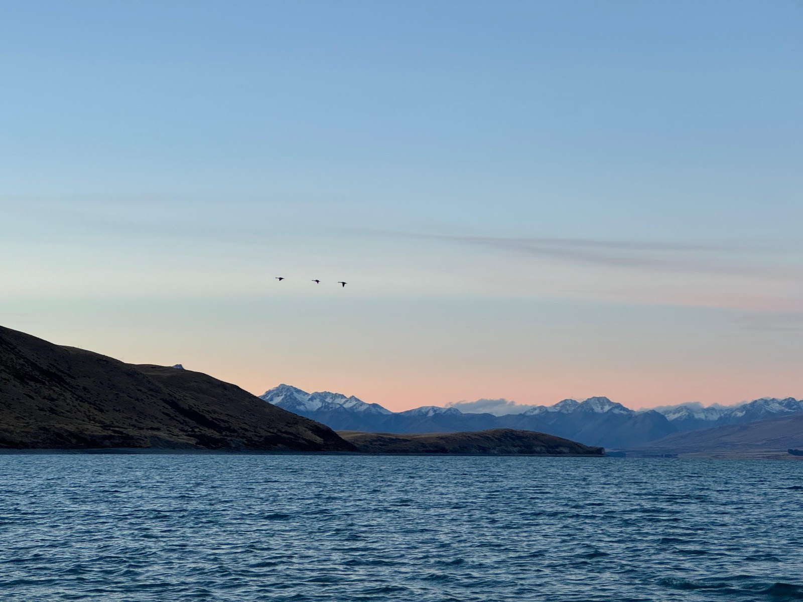 Still water at dusk with low hills beyond and three birds crossing a pale sky
